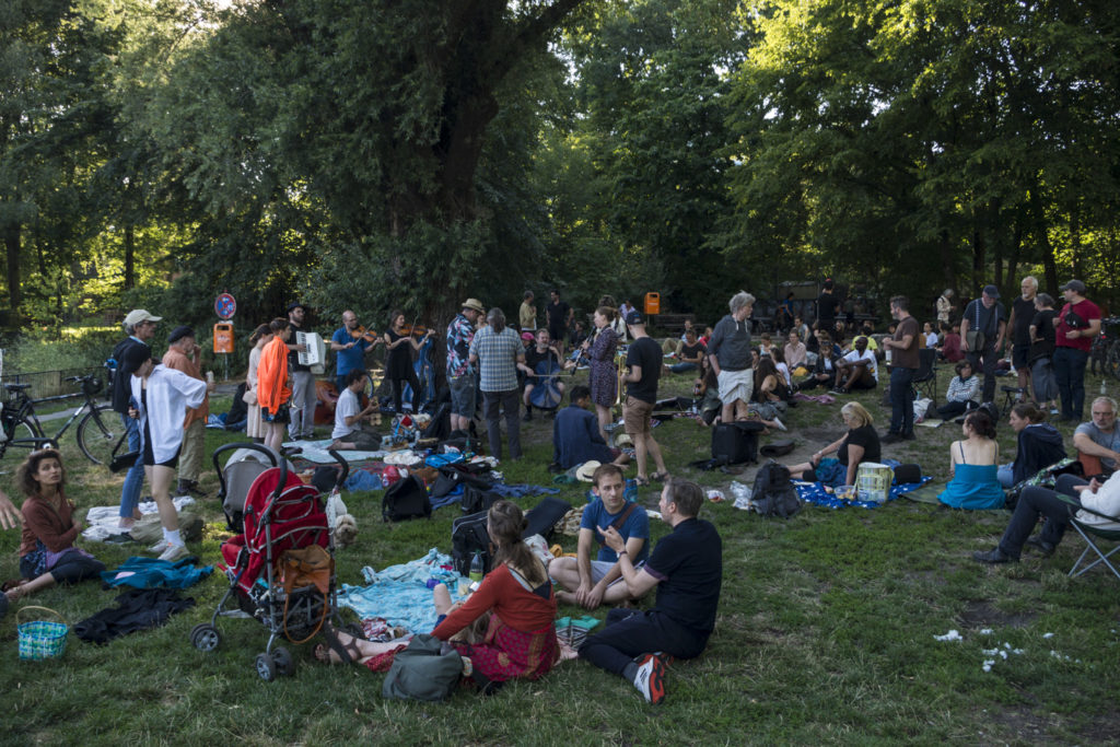 A photograph of the kleznick (klezmer picnic) in July, showing several dozen people sitting on picnic blankets, playing instruments including fiddles and accordions, and chatting in a grassy, leafy setting